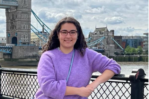 Photo of Kaita Gurian, a white woman with long curly hair and glasses wearing a purple sweater and blue satchel bag. She is standing in front of the Thames River and Tower Bridge in London