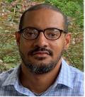 Dark skinned man with short hair and glasses looking at the camera