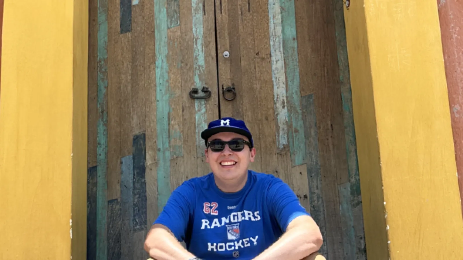 Man in blue shirt and ball cap sitting on the steps of a yellow doorway