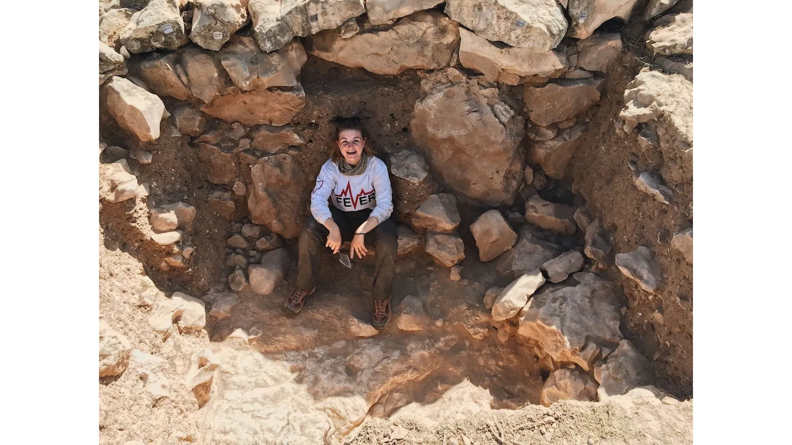 A young woman in a white top and brown pants sitting in an archaeological field site
