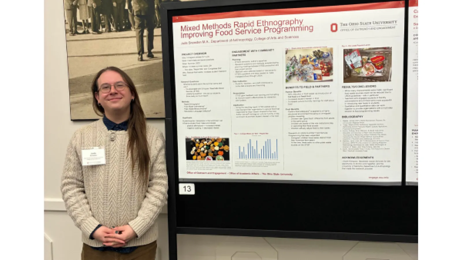 Young man in a brown sweater standing next to a poster at a conference
