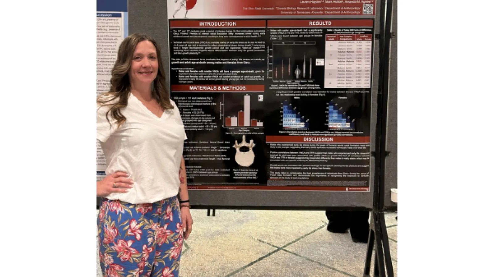A young woman with brown hear, wearing a white top and floral pants standing in front of a poster at a conference