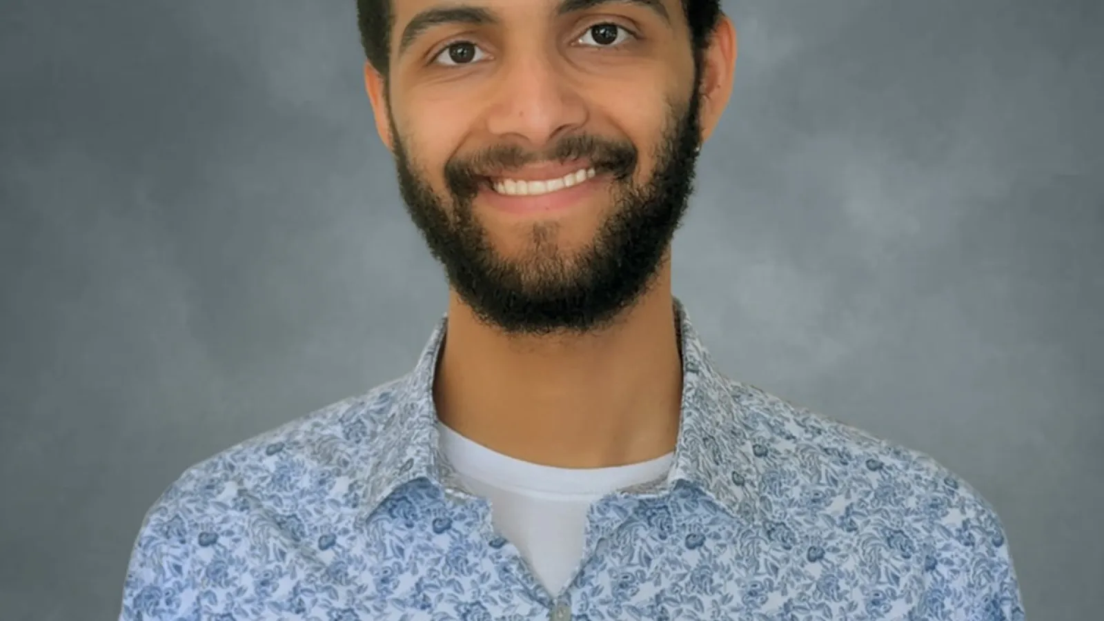 A young man with short dark hair and dark facial hair wearing a blue button down