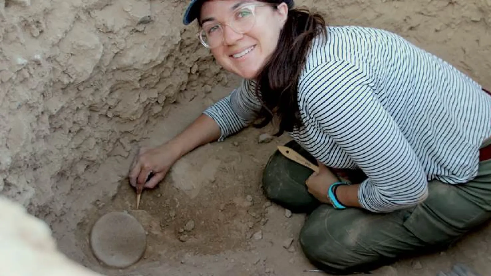 Brown haired woman in cap looking up from archaeological pit