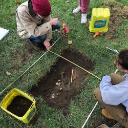ANTHROP5686 This image shows two students over a small "grave" that they excavated for their class ANTHROP5686. They are planning the location of several pieces of evidence, including plaster casts of bones.