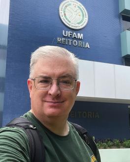 portrait of Butch Wright. Butch is a fair-skinned man with white hair. in this image he is wearing glasses, a green t-shirt and stand in front of a blue building, the UFAM, or Federal University of Amazonas