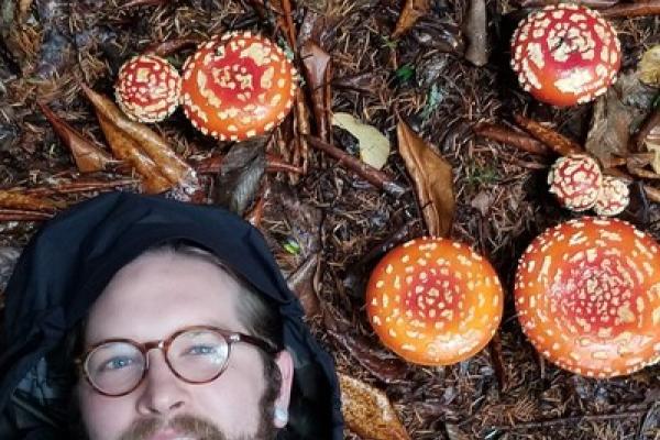Shane Scaggs; he is a white man with glasses and a beard laying on the ground next to some red and white mushroom caps.