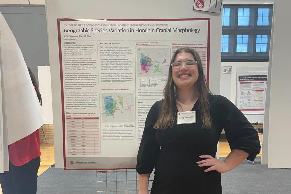 Photo of Abby Scheeser presenting a research poster. She is a fair-skinned woman wearing a black dress with long sleeves and clear thick-rimmed glasses