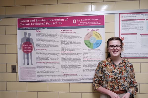 Picture of Madelin Zi Ching in front of her research poster hanging on a yellow tiled wall. she is wearing a floral blouse and khakis and has glasses and a ponytail