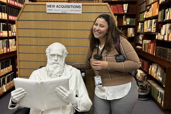 Grace Peters photo. Grace is wearing a brown sweater over a white dress shirt and black and white dotted slacks. She is in a library posing in front of a white statue of Charles Darwin.