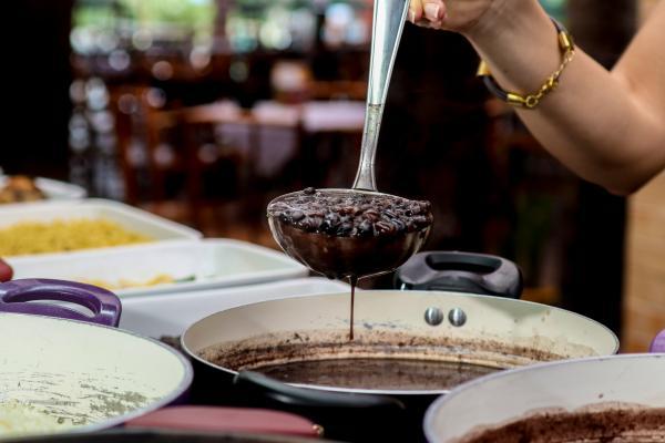 A ladle full of black beans being served out of a pot by a human arm with a gold chain