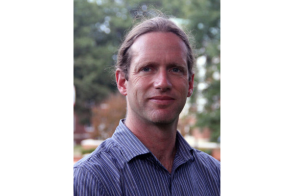 Dr. Sean Downey, a white man standing in front of a tree wearing a blue striped dress shirt