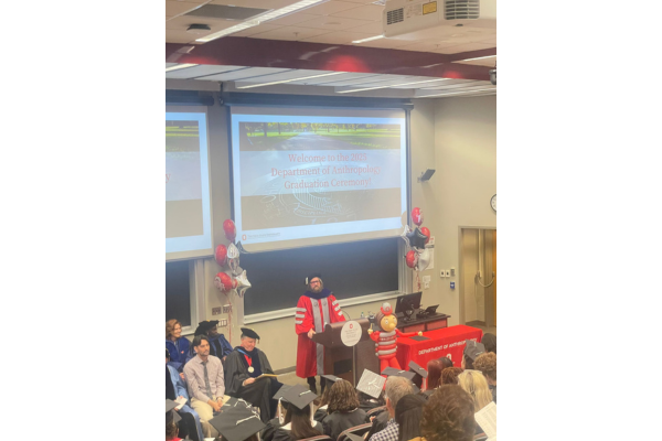 A young man wearing a scarlet and grey graduation robe giving a speech in an auditorium