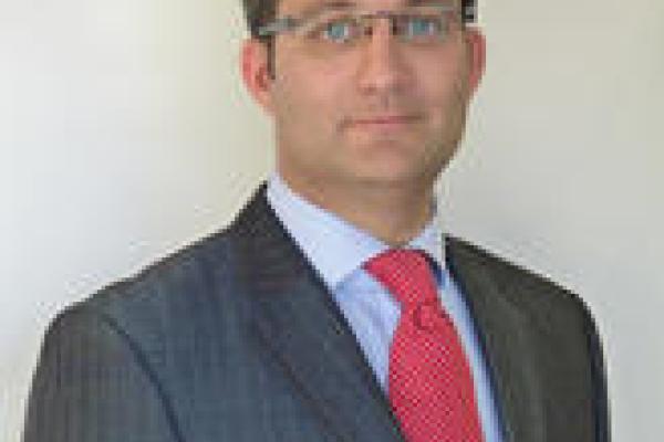 A brown haired blue eyed man in glasses with a red tie and blue sport coat