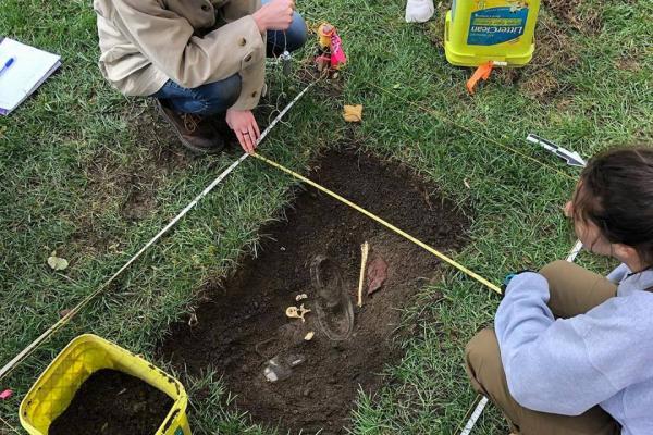 This image shows two students over a small "grave" that they excavated for their class ANTHROP5686. They are planning the location of several pieces of evidence, including plaster casts of bones. 
