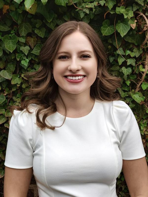 Allyson Simon; Allyson is a white woman with brown hair , in this image she is standing in front of green ivy and wearing a white short-sleeve shirt