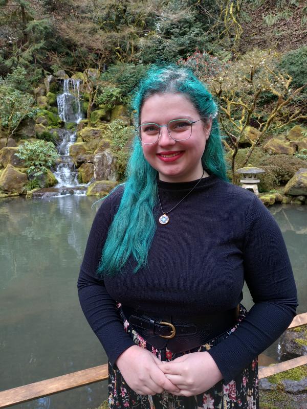 Julie Lierenz; white woman with blue hair and glasses, in the image she is wearing a navy sweater, floral skirt and standing in front of a waterful