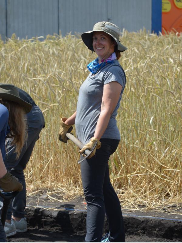 Lauren Hayden; in this image Lauren is in the field holding a pickaxe and wearing a bucket hat, behind he is tall grass. she is wearing black jeans and a gray t-shirt