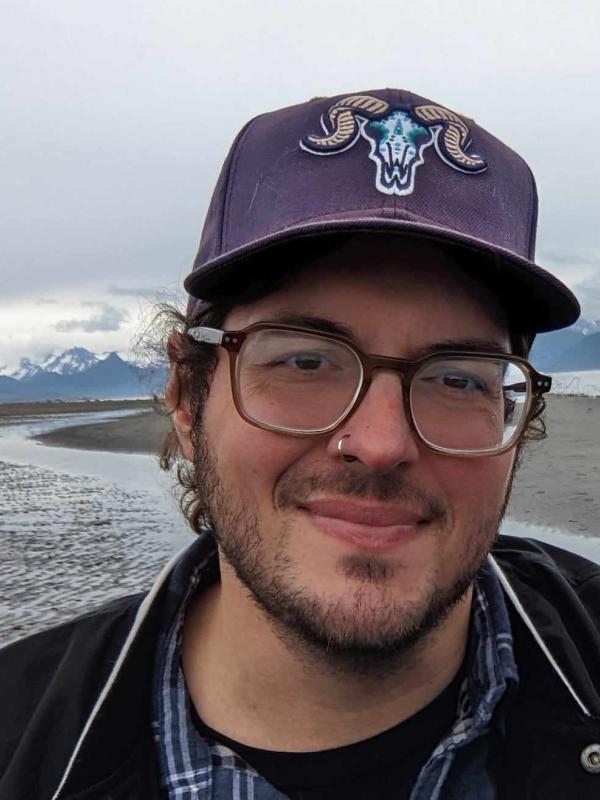 Nick Mauro; Nick is a white man with glasses and a beard; in this iage Nick is wearing a purple baseball gap and sending in front of a glacier