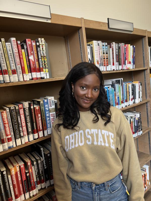 Nina Wilson; Nina is a Black woman with black hair, in this image she is wearing a beige Ohio State sweatshirt and jeans and standing in front of two bookcases