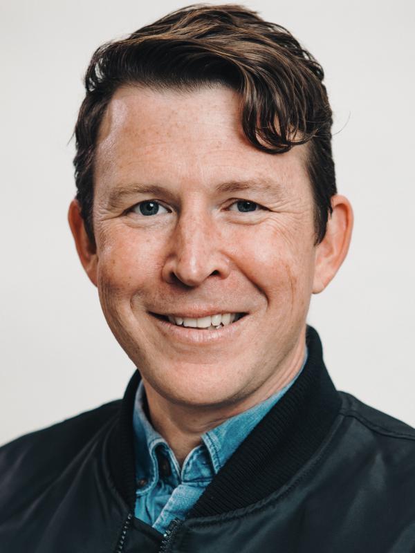 Photo of a white man with short brown hair, a blue collared shirt, and a black bomber jacket
