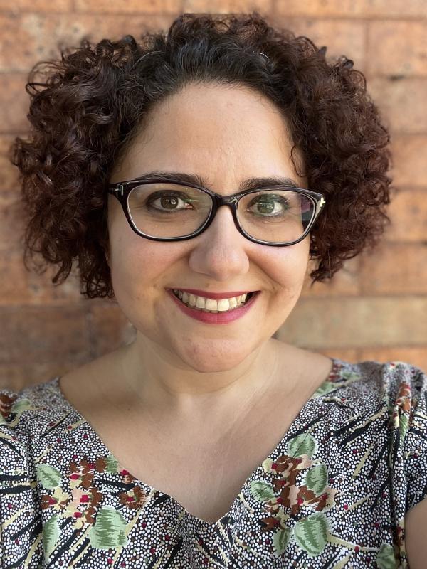 Bürge is a woman with short curly brown hair and glasses. in this image she is smiling and standing in front of a brick wall, wearing a black and white blouse