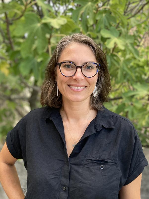 photo of Céline Lamb; she is a white woman with short brown hair and glasses. in this image she is standing in front of a tree and wearing a black blouse