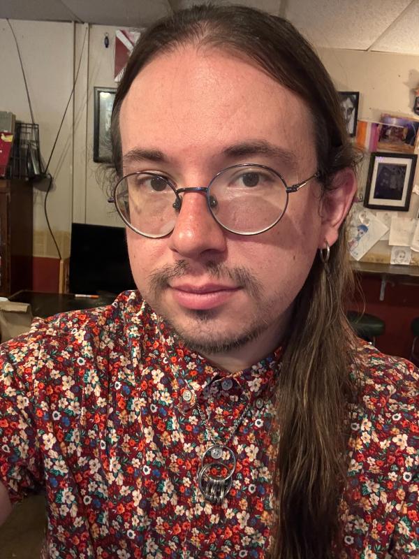 Jude Snowden, white with long brown hair and a goatee, round glasses, in a floral print shirt with a dark red background and silver hoop earrings