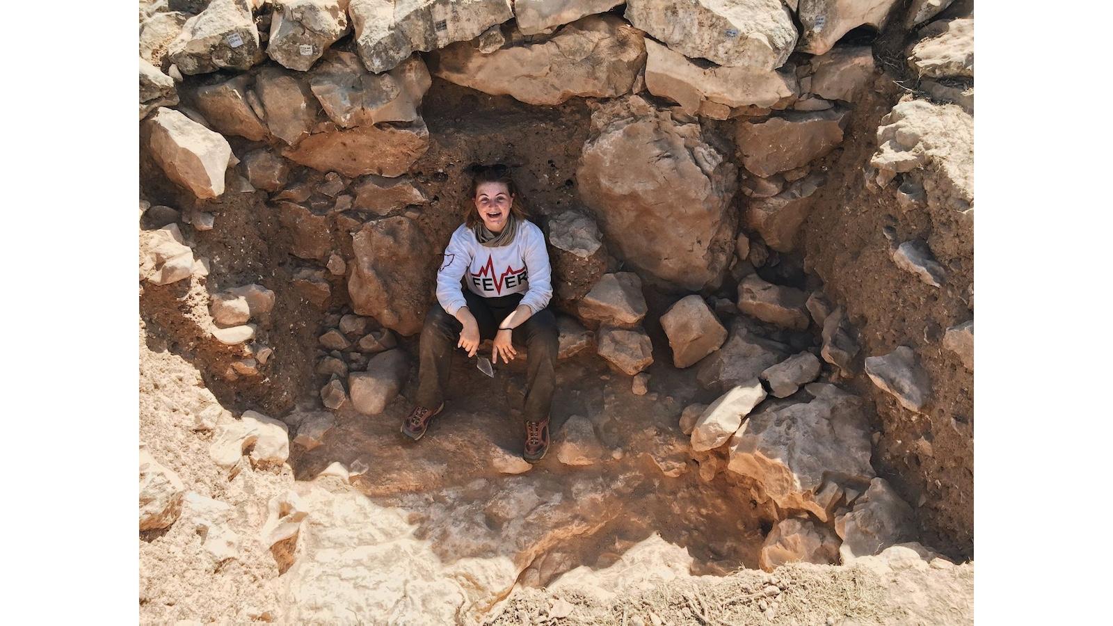 A young woman in a white top and brown pants sitting in an archaeological field site