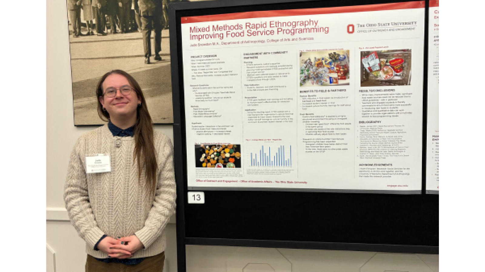 Young man in a brown sweater standing next to a poster at a conference