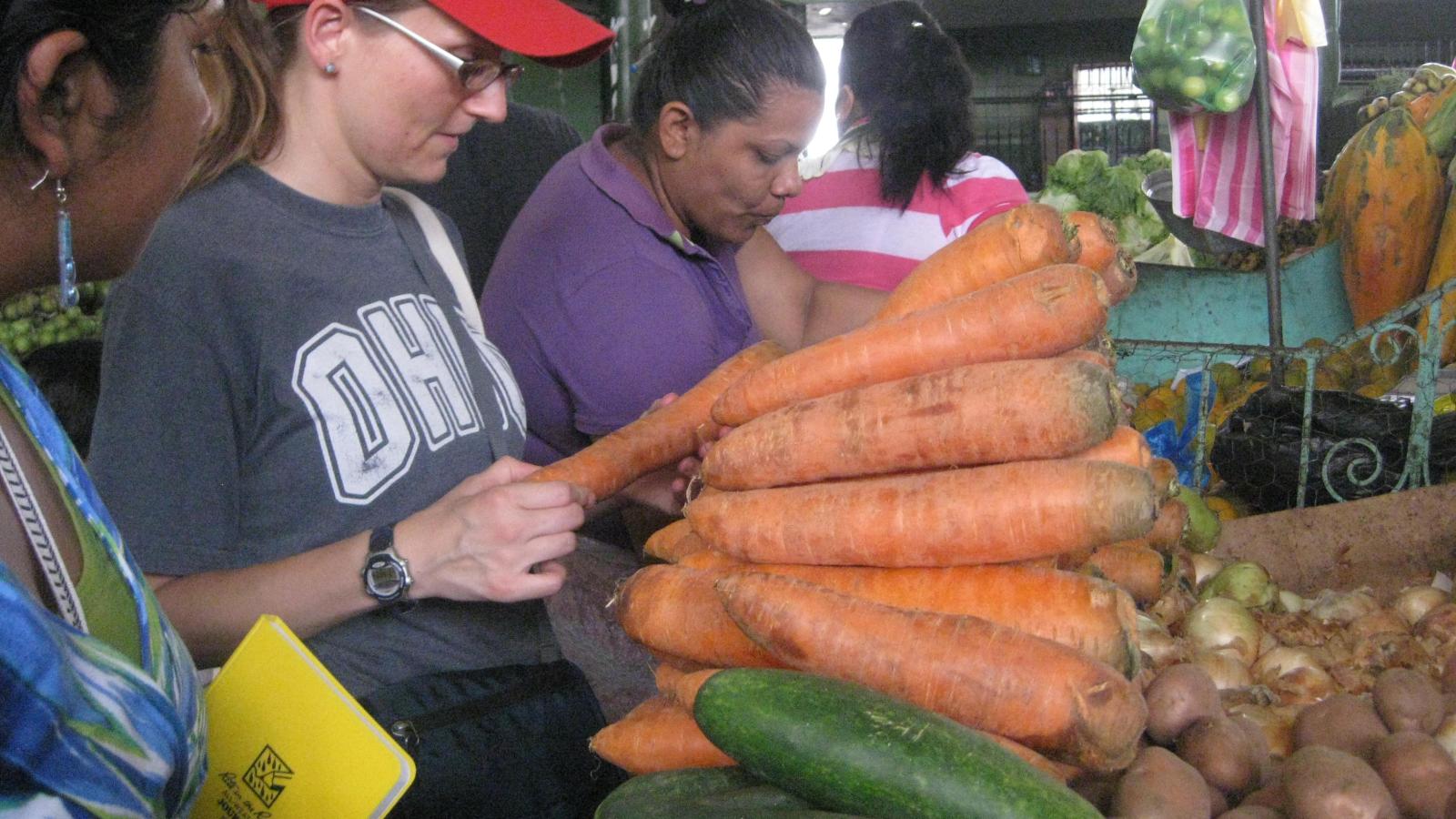 student in red ball cap inspecting vegetables at a market
